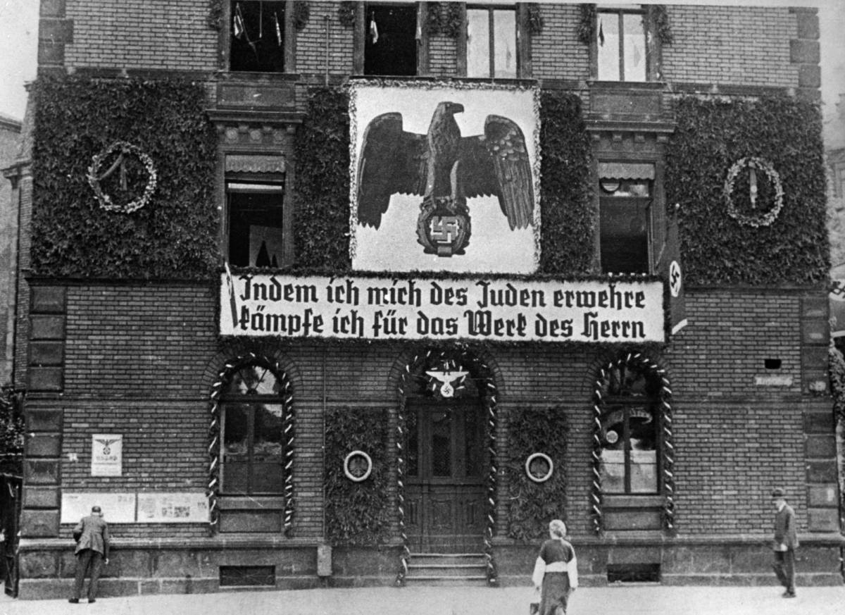 An antisemitic message hangs outside a Nazi headquarters. The banner reads, ‘By resisting the Jew, I fight for the work of the Lord.’