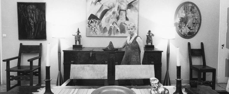 Peggy Guggenheim in the dining room of Palazzo Venier dei Leoni, Venice, 1960s. On the wall (left to right) Marcel Duchamp, Nude (Study), Sad Young Man on a Train (1911-12 PGC), Vasily Kandinsky, Landscape with Red Spots, No. 2 (1913, PGC), Georges Braque, The Clarinet (1912, PGC); in the foreground, on the table, Jean Arp, Head and Shell (1933, PGC).
