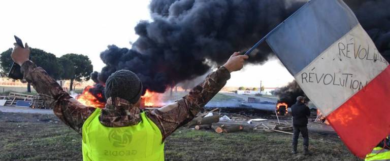 A man wearing a yellow vest waves a French flag in front of burning tires at a road blockade during a protest against the rise in the price of oil and the cost of living, on Dec. 11, 2018, in southern France. 