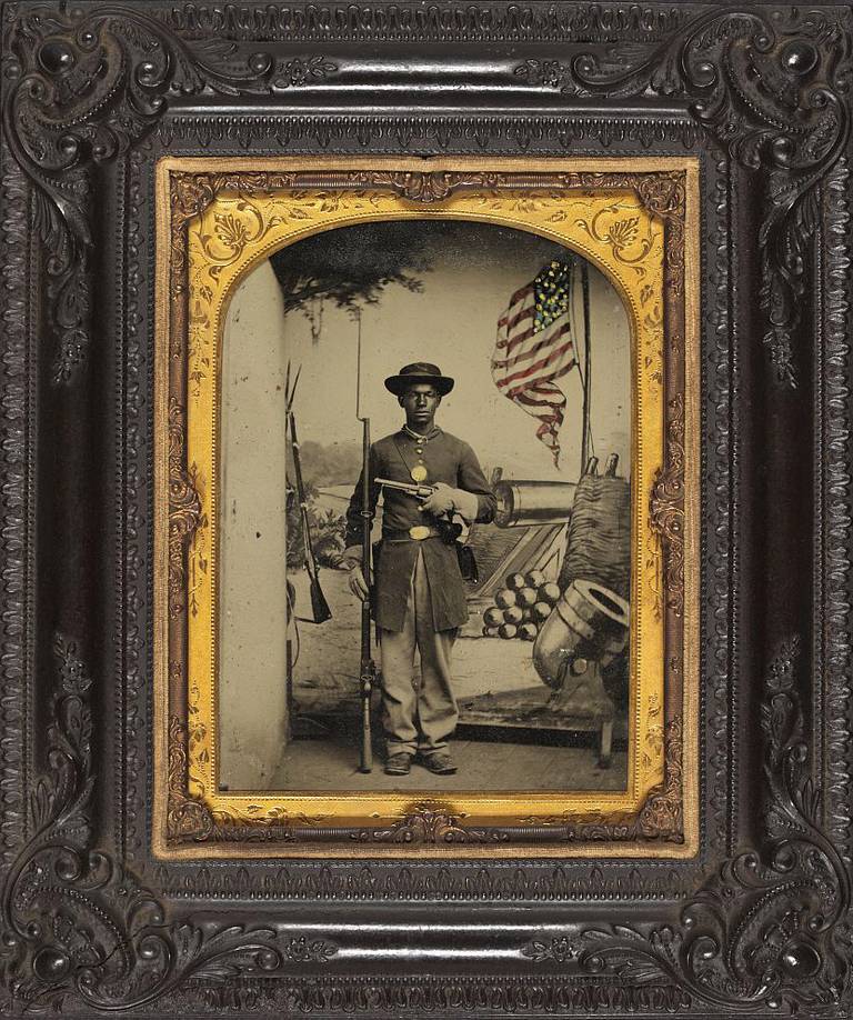 Unidentified African American soldier in Union uniform with a rifle and revolver in front of painted backdrop showing weapons and American flag at Benton Barracks, Saint Louis, Missouri.