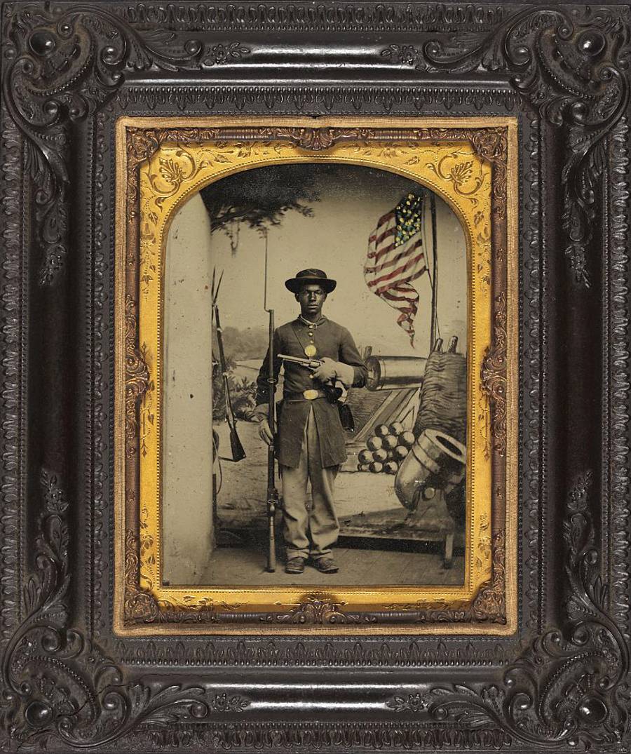 Unidentified African American soldier in Union uniform with a rifle and revolver in front of painted backdrop showing weapons and American flag at Benton Barracks, Saint Louis, Missouri.