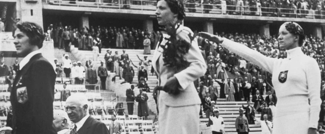 The three winners of the Women's Individual Foil competition in the 1936 Olympic Games stand on the podium in Berlin as spectators applaud. From left: bronze medalist Ellen Preis of Austria, gold medalist Ilona Elek of Hungary, and silver medalist Helene Mayer of Germany. 
