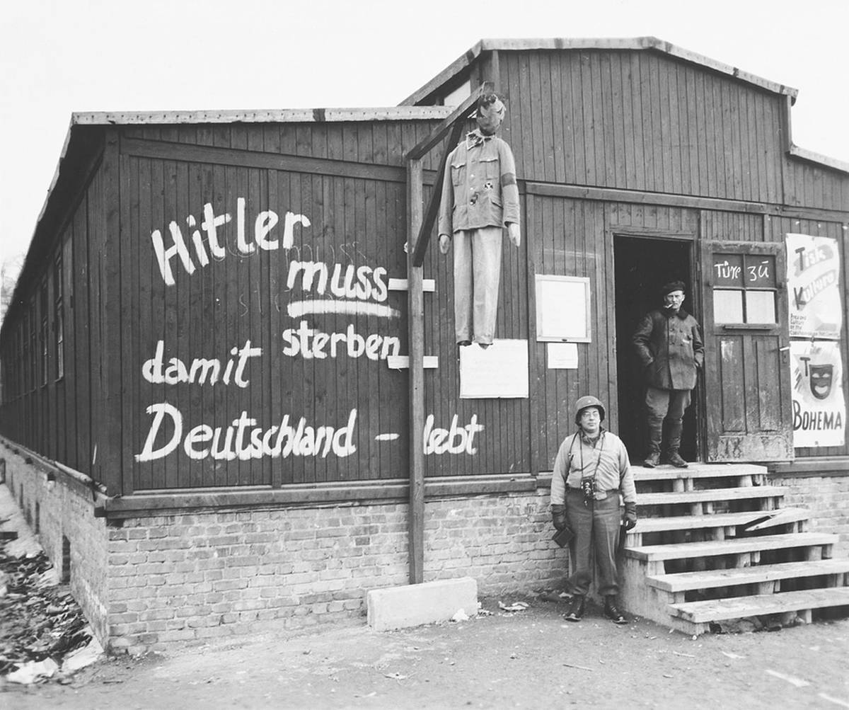 An American soldier poses next to an effigy of Hitler hanging in front of a barracks in the Buchenwald concentration camp. Graffiti on the side of a barracks reads, ‘Hitler must die for Germany to live,’ 1945.
