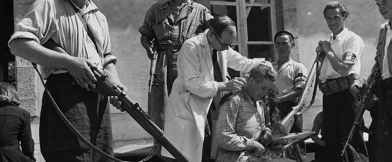 A barber shaves the head of a woman accused of being a Nazi collaborator, surrounded by members of the Free French Resistance with rifles, Bourg-Blanc, France, 1944